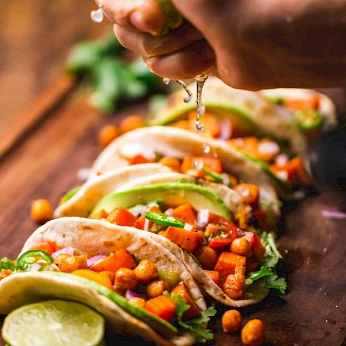 A hand squeezes lime juice over vibrant, fresh veggie tacos with chickpeas, avocado, tomatoes, and cilantro, all neatly arranged on a wooden board.