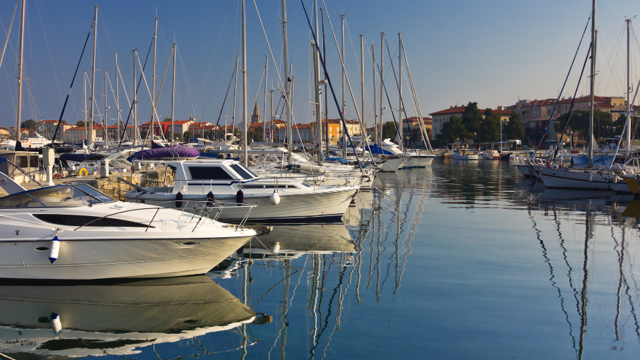 A marina with several boats docked, calm water reflecting the boats and clear sky, with buildings in the background ending the sentence.