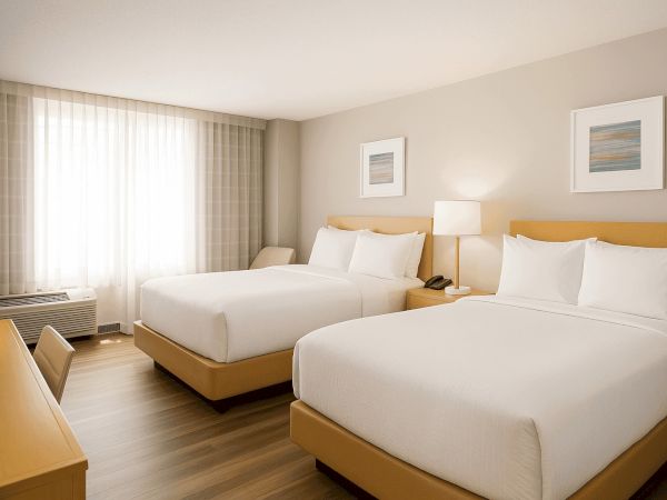 The image shows a hotel room with two neatly made double beds, wooden furniture, a lamp, and framed artworks on the wall.
