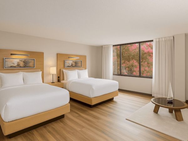 The image shows a hotel room with two beds, paintings above them, a window with curtains and a view, and a table with a decorative item.