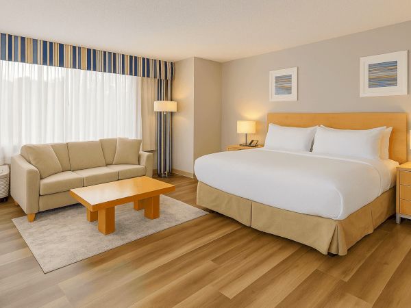 The image shows a hotel room with a large bed, beige sofa, coffee table, side tables, lamps, and artwork on the wall, with wooden floors.