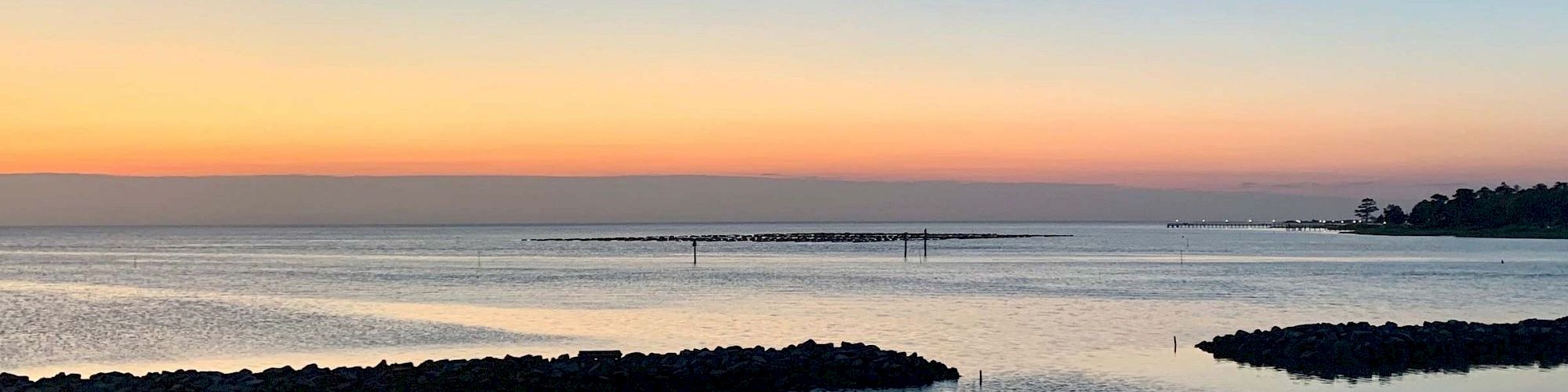 A tranquil coastal scene at sunset: calm water with rock breakwaters, a soft gradient sky from orange to blue, distant shoreline, and a peaceful horizon.